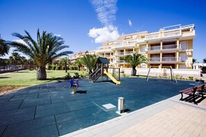 A playground with a slide and swings, surrounded by palm trees and a building with balconies. - 176 Estrella de Mar, 6 (denia)