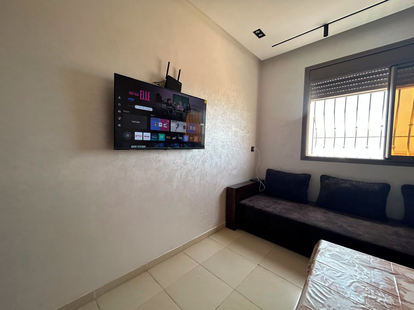 A room with a flat-screen TV mounted on the wall displaying various streaming services, a sofa with cushions, a window with blinds, and a tiled floor.