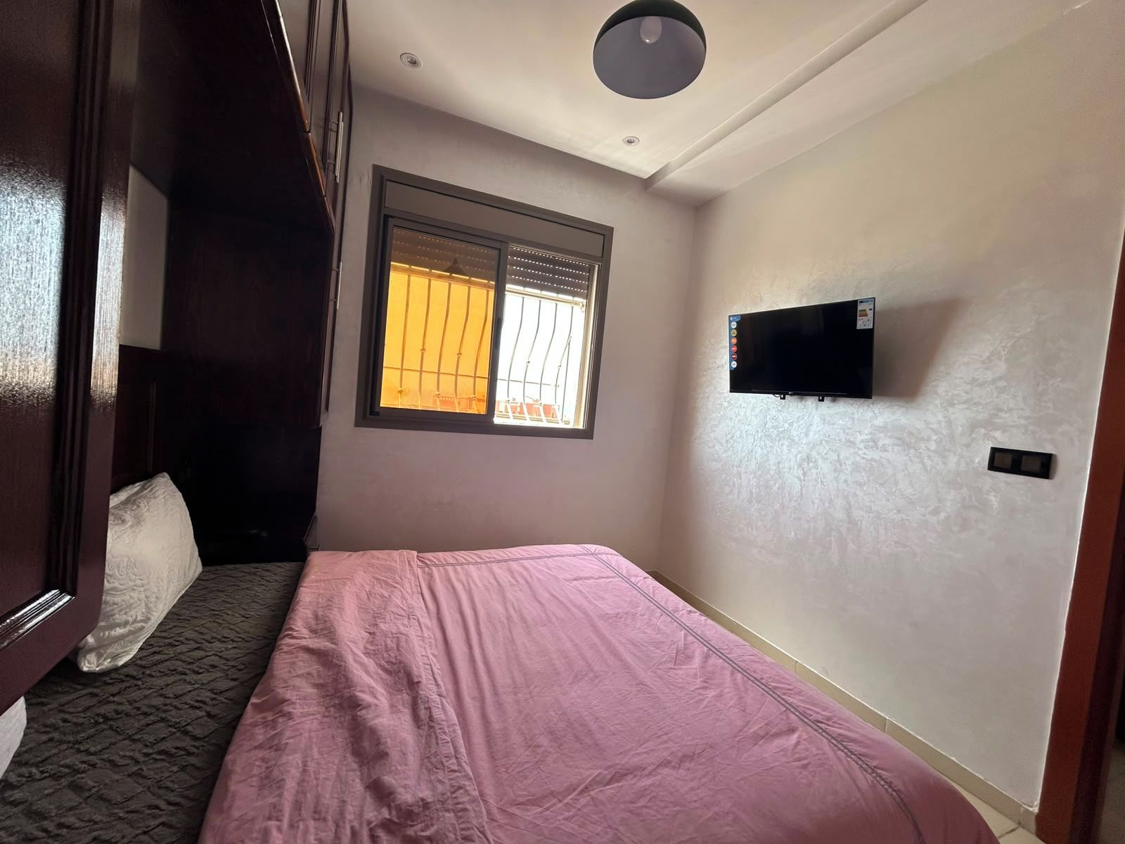 A bedroom with a bed, a window with blinds, a flat-screen TV mounted on the wall, and a ceiling light fixture.