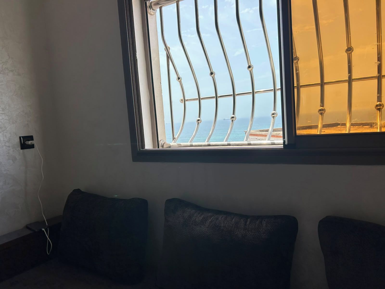 A room with a window overlooking a body of water, a power outlet, and two dark-colored cushions on a low table.