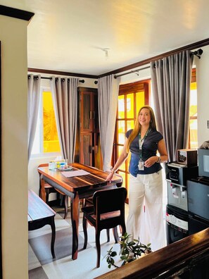 Una persona de pie en una habitación con una mesa de comedor de madera, sillas, un refrigerador y una ventana con cortinas.