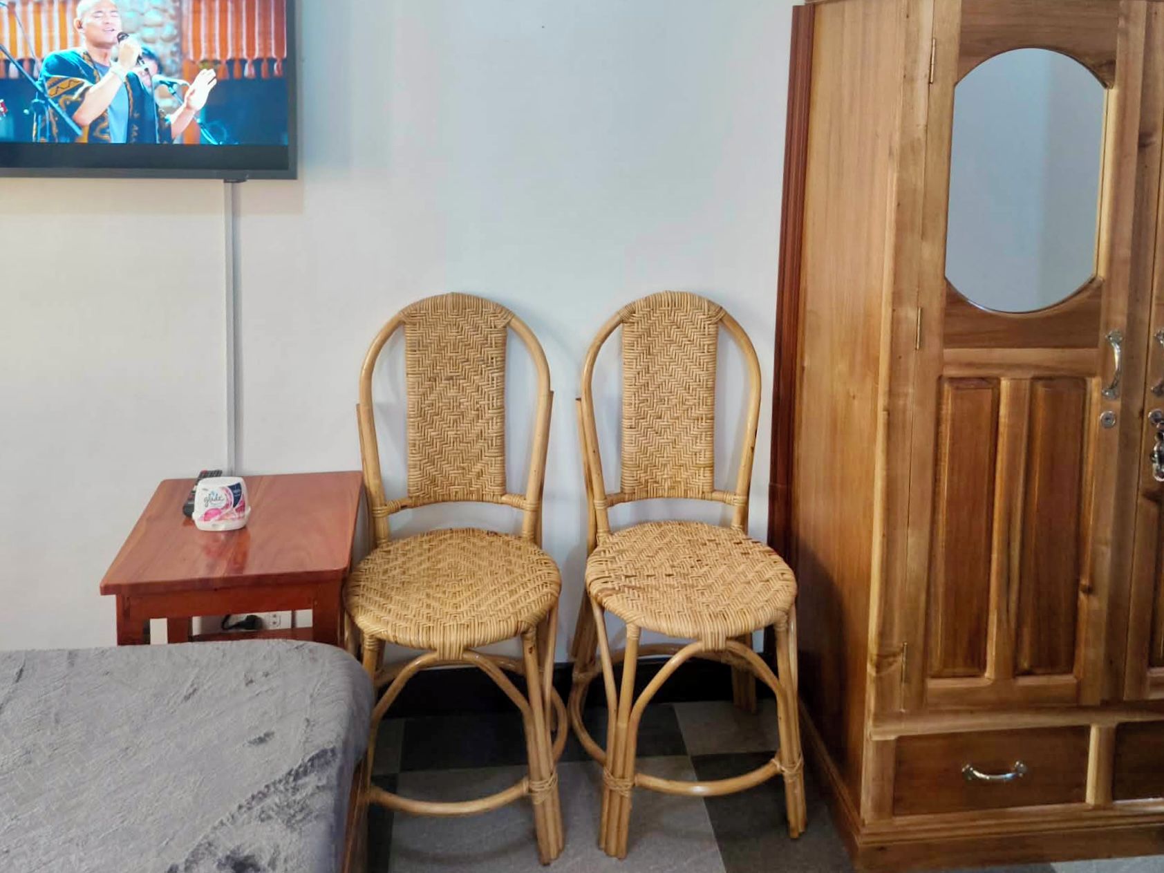 Une pièce comprenant deux fauteuils en osier, une petite table avec un coffre, une armoire en bois et un téléviseur fixé au mur.