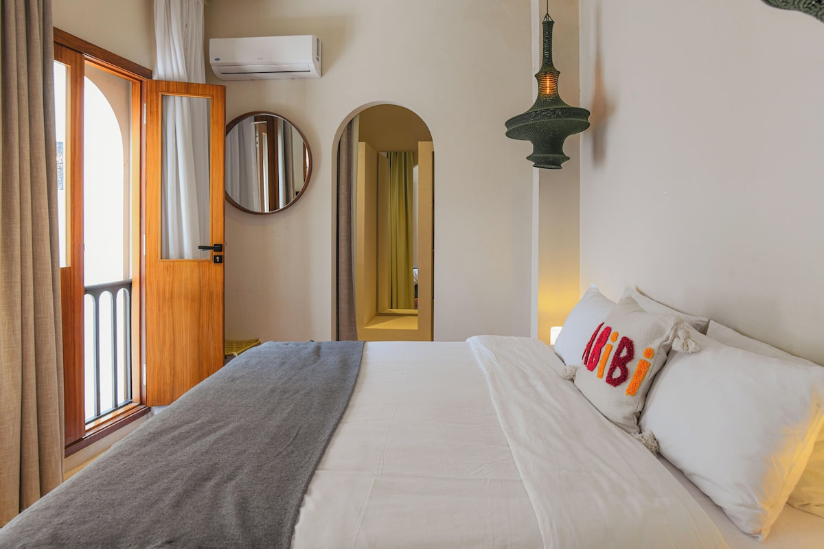 A hotel room with a bed, a mirror, an air conditioning unit, a door, and a hallway.