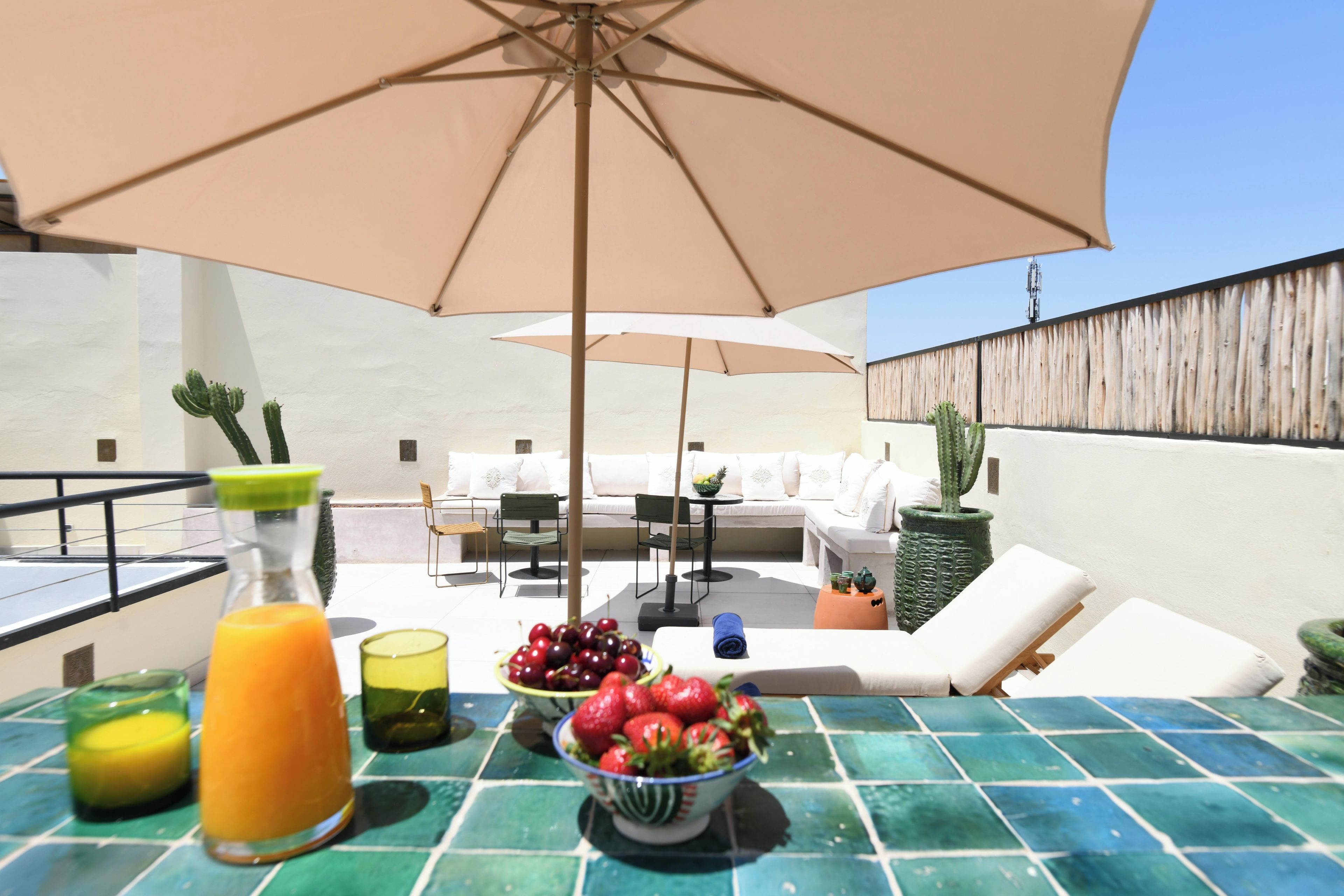 A rooftop terrace with outdoor seating, a table with fruit and drinks, and a large umbrella providing shade.