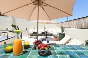 A rooftop terrace with outdoor seating, a table with fruit and drinks, and a large umbrella providing shade. - Asilah (Marrakech)