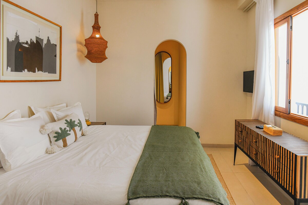 A neatly arranged bedroom with a bed, pillows, a hanging light fixture, a mirror, a bedside table, and a window with curtains.