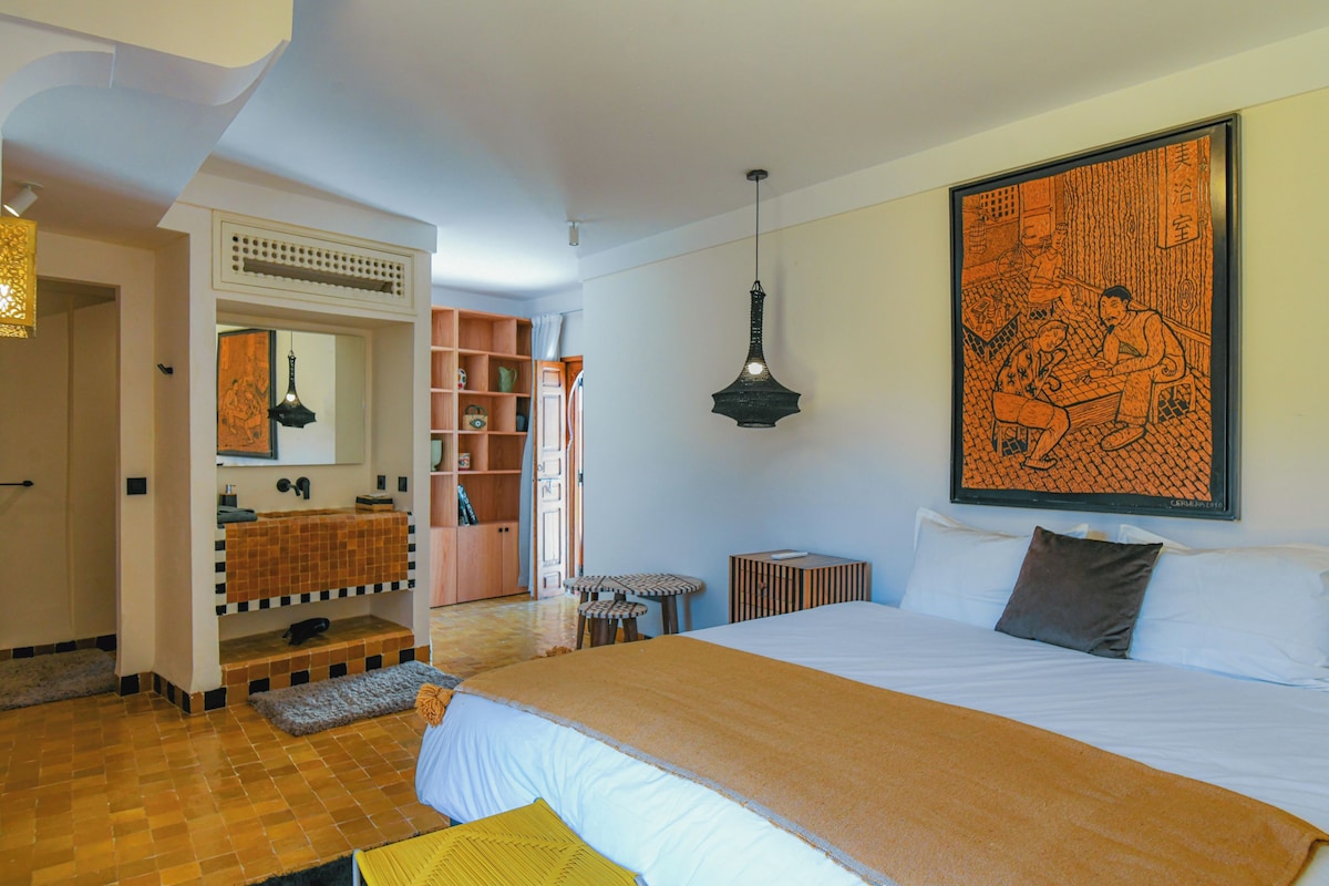 A hotel room with a large bed, a bathroom with a sink and mirror, a bookshelf, and a large artwork on the wall.