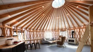Interior de una yurta con una cúpula central de madera, un sofá, una mesa de comedor y una zona de cocina.