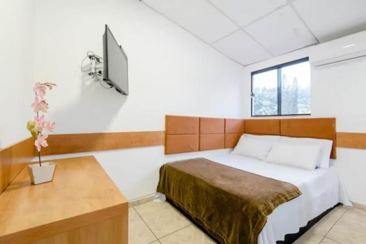 A hotel room with a bed, a TV mounted on the wall, a window, and a small table with a vase of flowers.