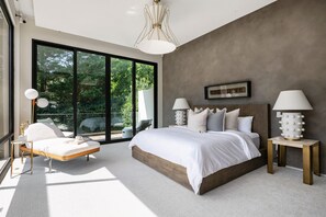 Une chambre moderne avec un grand lit, deux tables de chevet avec des lampes, une chaise longue et une vue sur la verdure grâce à de grandes fenêtres.