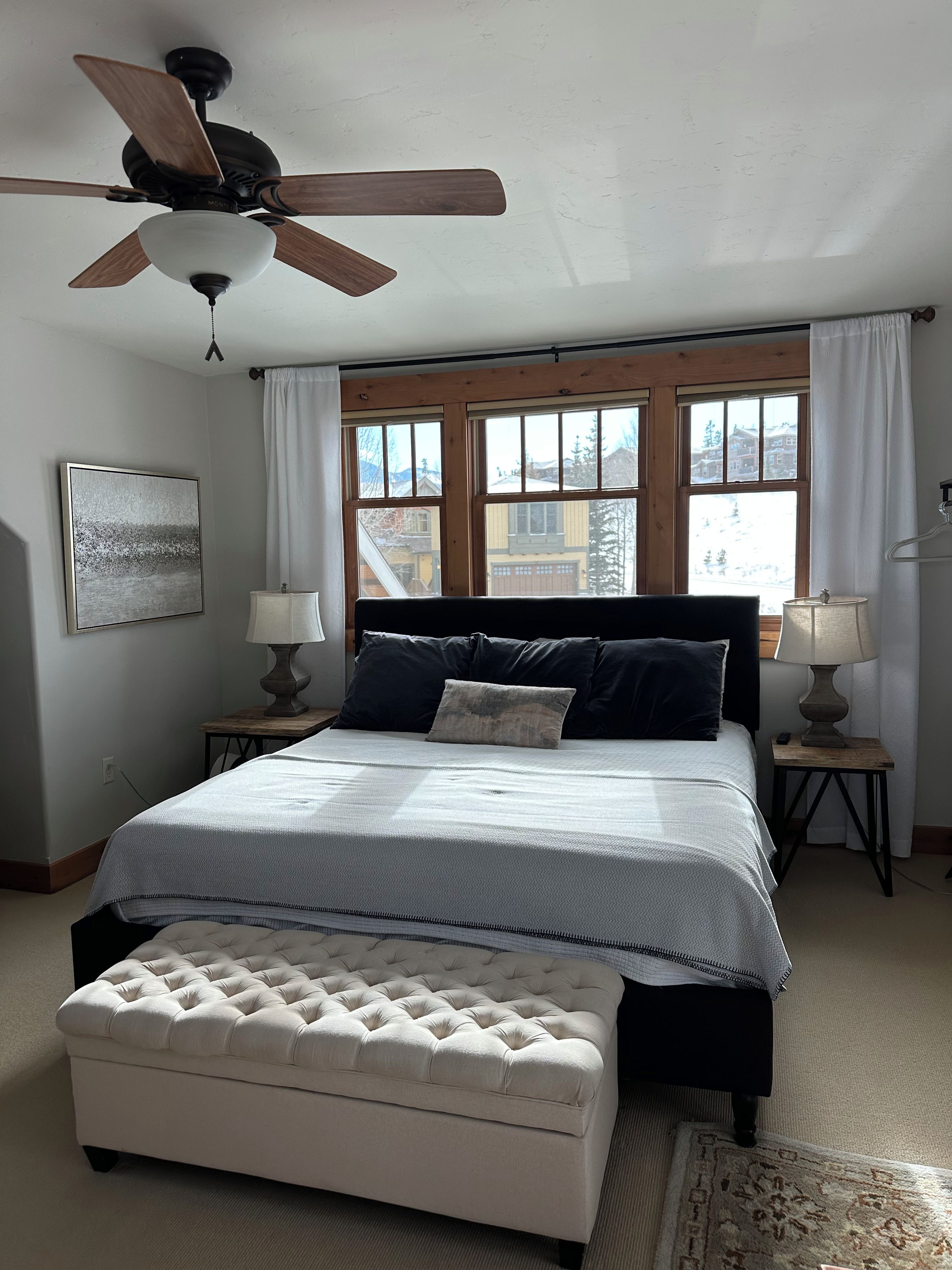 A bedroom with a bed, a ceiling fan, two bedside tables with lamps, a window with curtains, and a rug on the floor.