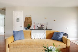 Ein Wohnzimmer mit einer Couch, Kissen, einer Tür, einem Wandspiegel, einem kleinen Tisch mit Dekoration und einer Pflanze.