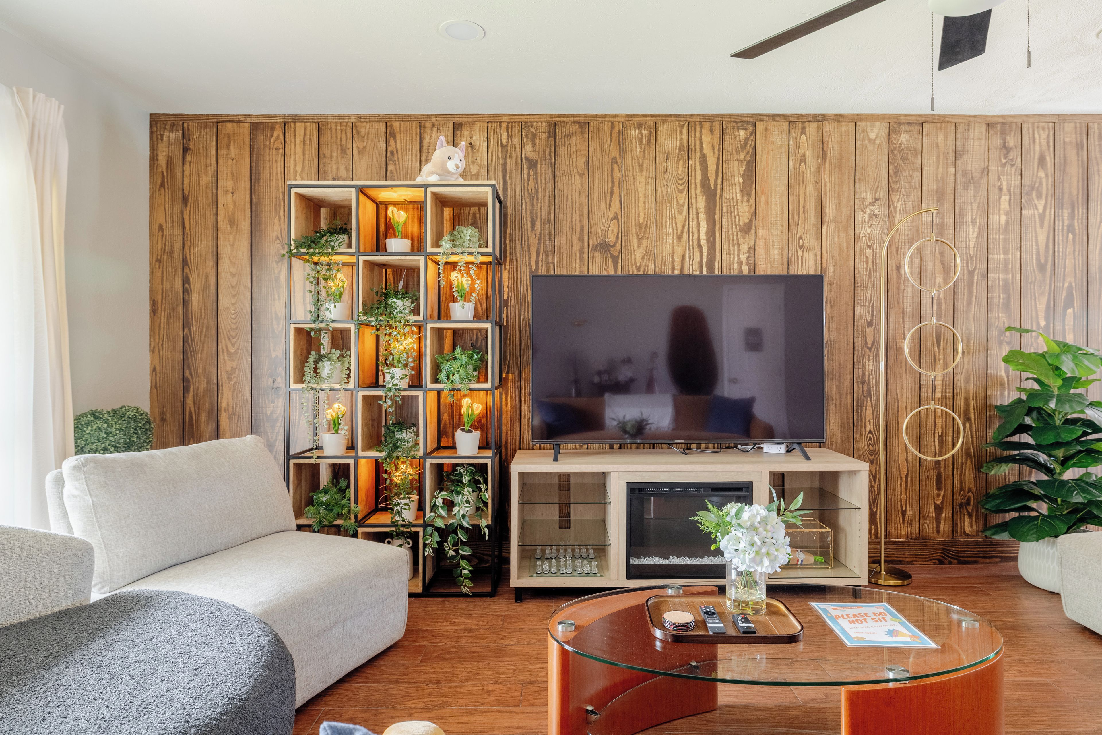 A modern living room with a wooden wall panel, a large flat-screen TV, a glass coffee table, and a sofa.