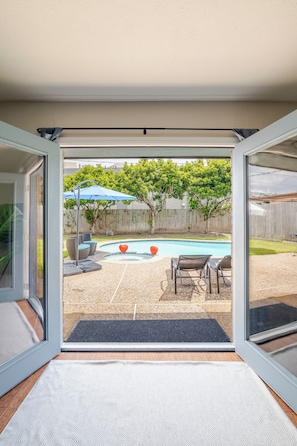 A view through a glass door into a backyard with a swimming pool, patio furniture, and a wooden fence. - Private Home with Jacuzzi and Large Deep Pool close to NRG/Galleria/Downtown (Houston)
