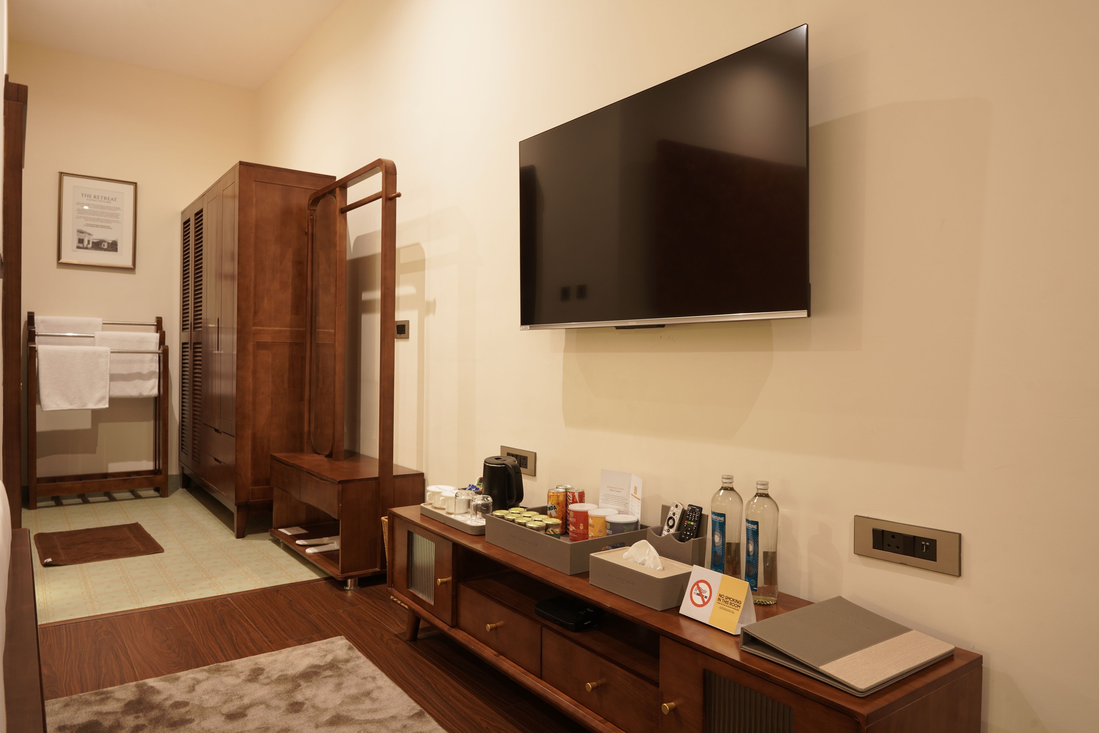 A hotel room with a flat-screen TV mounted on the wall, a wooden cabinet with drawers, a small table with toiletries, and a sign indicating no smoking.