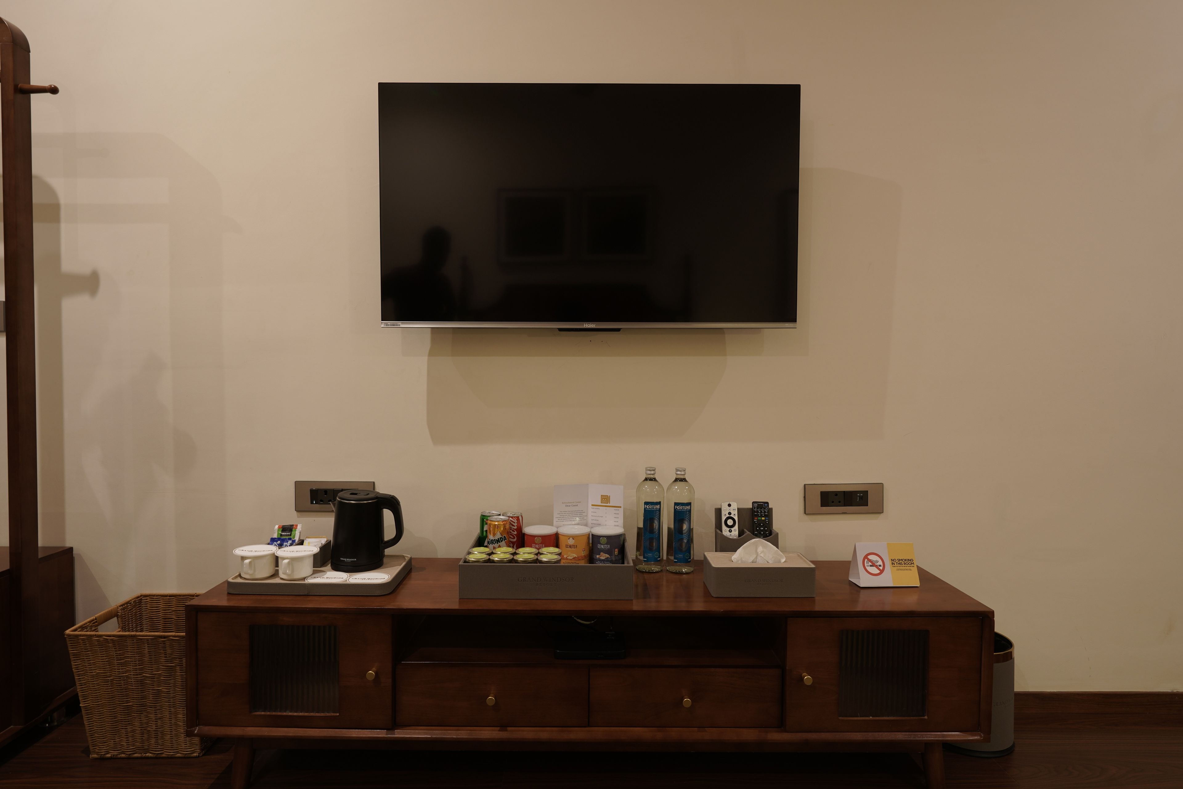 A hotel room with a flat-screen TV mounted on the wall, a wooden console table with a coffee maker, tea pots, and bottled water.