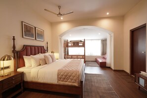 A hotel room with a large bed, a desk, a chair, a wardrobe, a ceiling fan, and a view of the outside. - Grand Windsor Resort (Ooty)