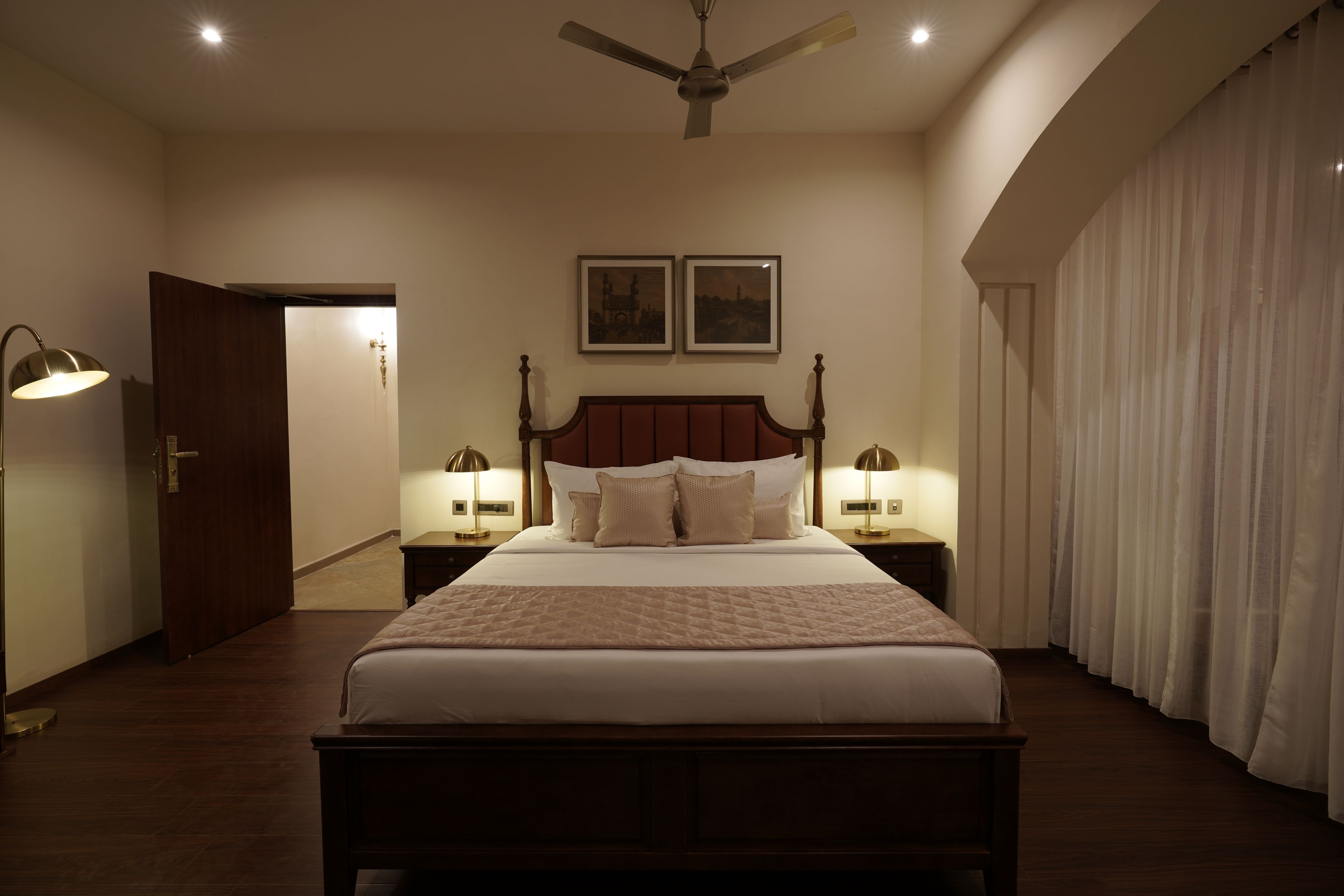 A bedroom with a large bed, two bedside tables with lamps, a ceiling fan, and two framed pictures on the wall.