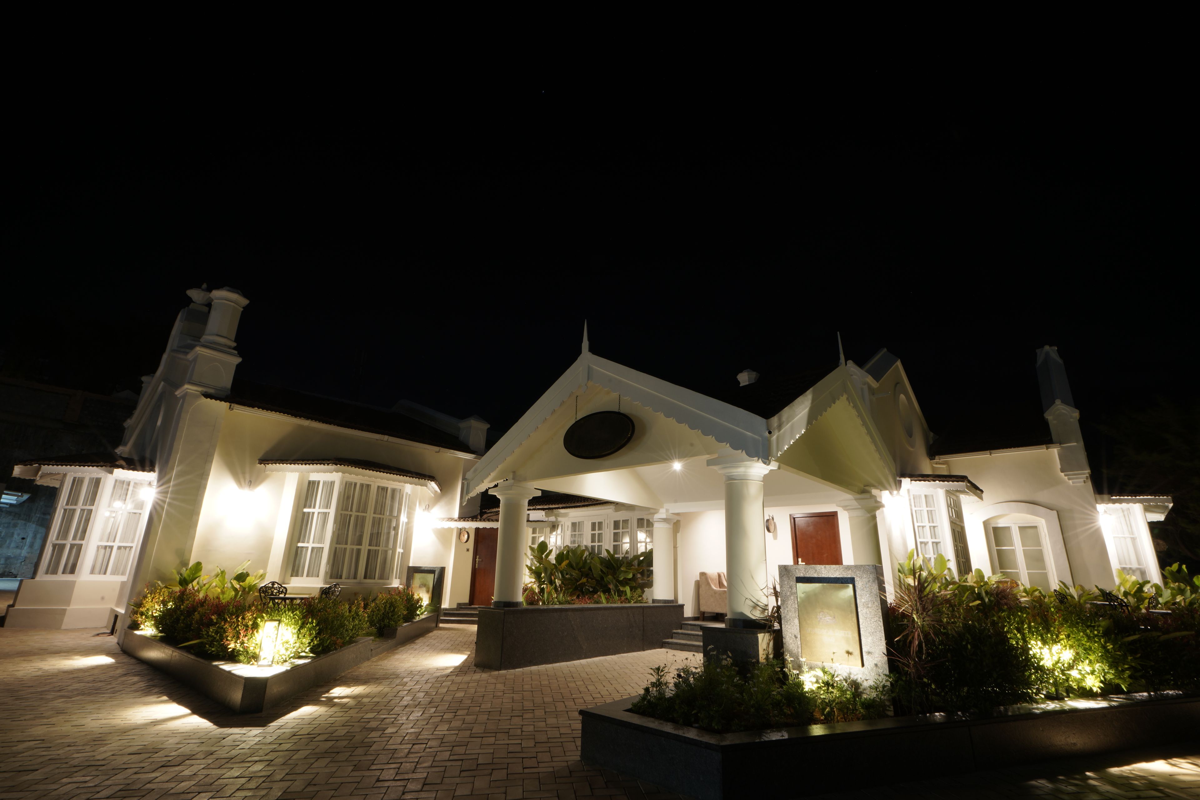 A well-lit, modern house with a brick pathway leading to the entrance, surrounded by lush greenery.