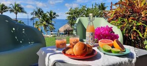 A table with a bottle, glasses, a plate with bread, and fruit, set against a backdrop of a beach and palm trees. - Turquoise Studio, Ocean View (Le Diamant)