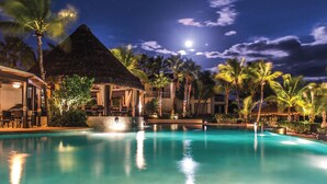 Zona de piscina nocturna en un resort, con un edificio de techo de paja, palmeras y cielo despejado.