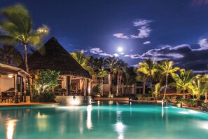 Área de piscina num resort à noite, com um edifício de telhado de palha, palmeiras e céu claro.