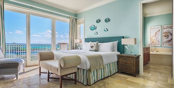 Quarto espaçoso com uma cama grande, uma cadeira, uma mesa de cabeceira e vista para o oceano através de grandes janelas.