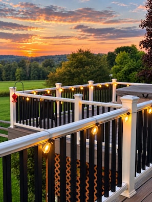 A deck with white railings and string lights, overlooking a green landscape during sunset.