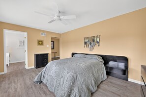 A bedroom with a bed, a ceiling fan, a wall-mounted artwork, and a door leading to another room.