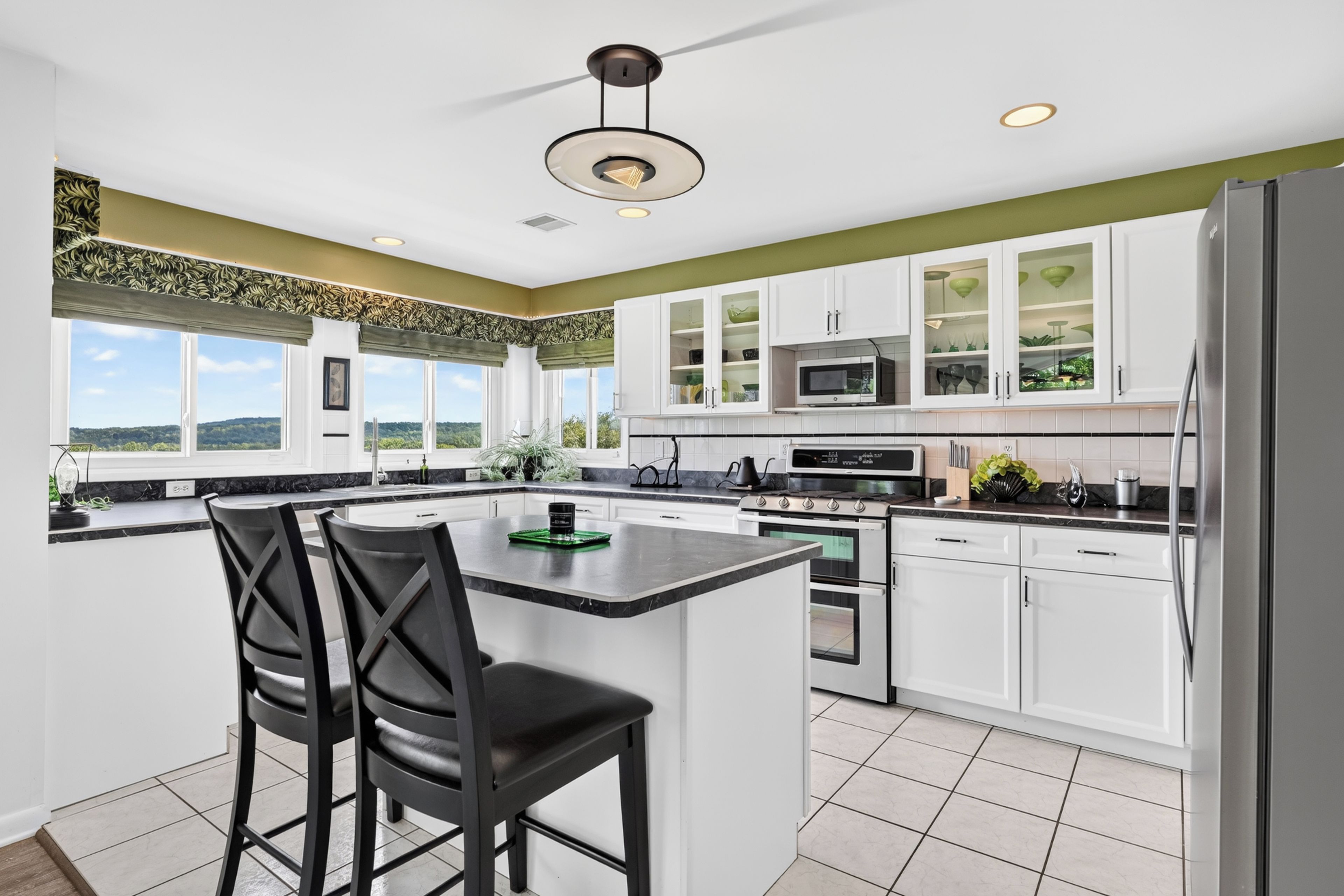 A modern kitchen with white cabinets, a central island with bar stools, a stainless steel refrigerator, and a view of the outdoors.