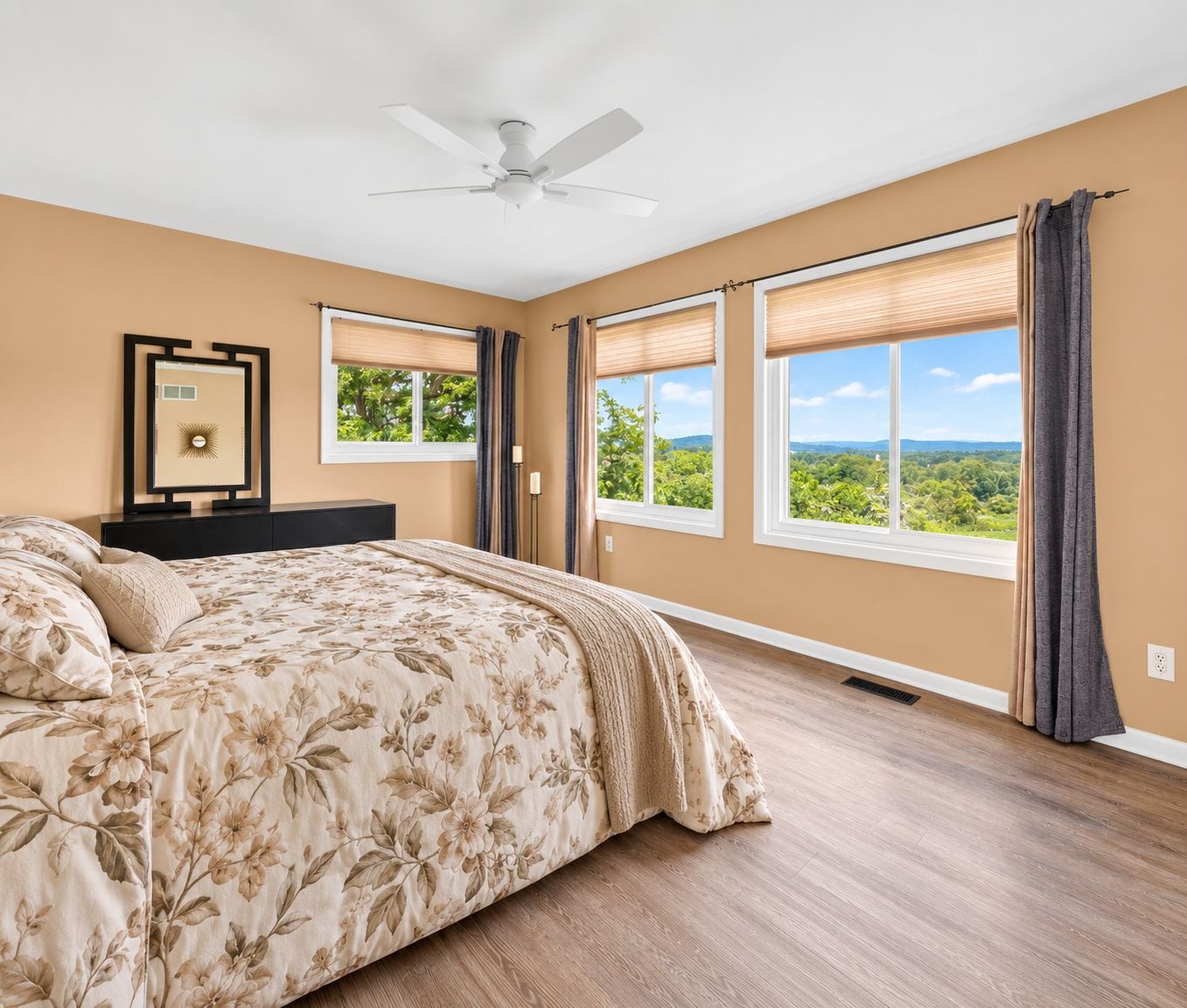 Ein Schlafzimmer mit Bett, Deckenventilator, großen Fenstern mit Jalousien und Blick auf Grünflächen.
