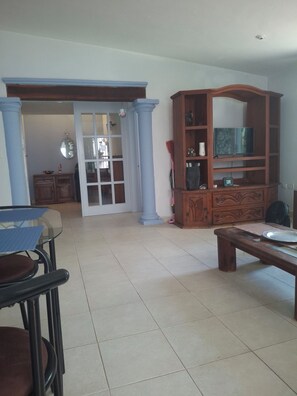 A living room with a glass table, a wooden bench, a television on a wooden cabinet, and a door leading to another room. - PURPLE LOTUS COZUMEL (Cozumel)