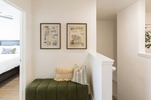 Une chambre à coucher avec un lit, un banc recouvert d’un tissu vert, deux tableaux abstraits encadrés et un escalier.