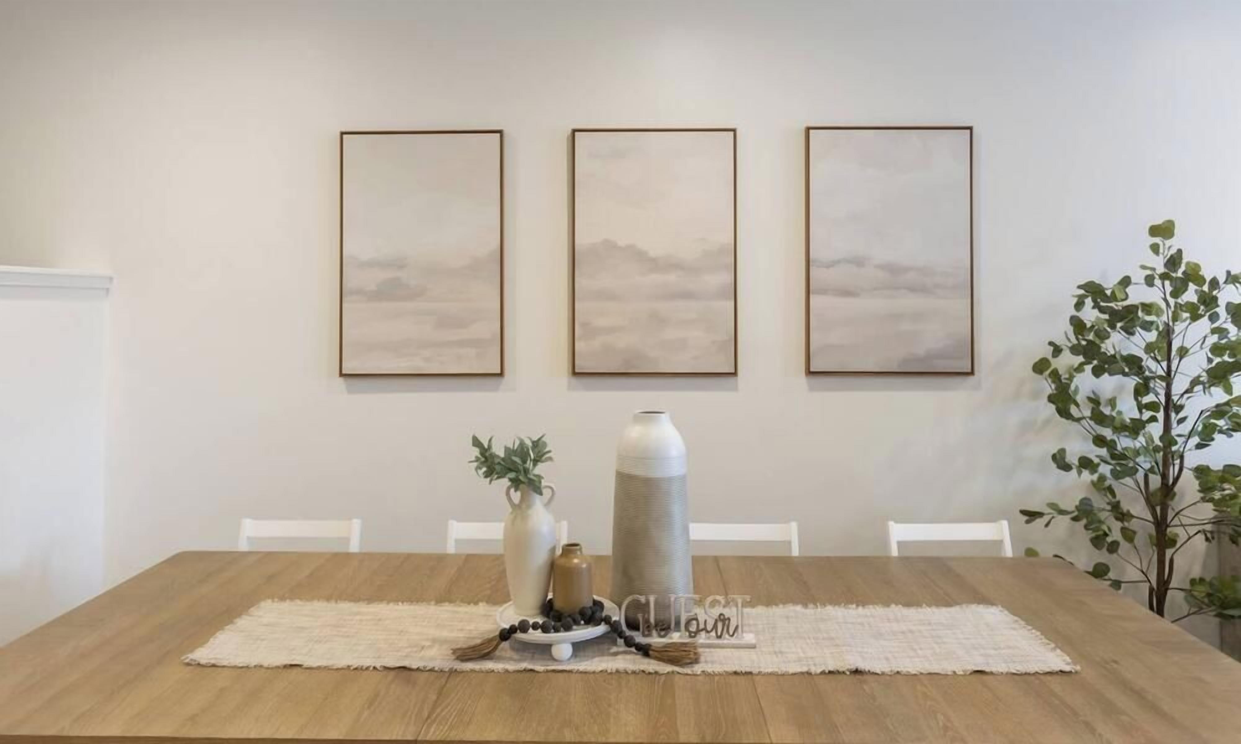 A dining table with a placemat, a vase with greenery, a tall vase, and a decorative item with berries. Three framed artworks are hung on the wall above the table.