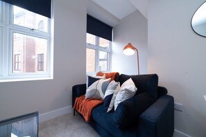 Ein modernes Wohnzimmer mit einem dunkelblauen Sofa, mehreren Kissen, einer Stehlampe und einem großen Fenster mit Blick auf ein Backsteingebäude.