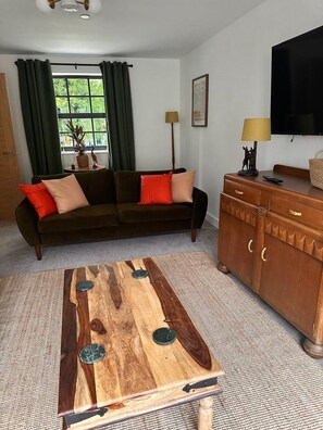 A living room with a sofa, a wooden coffee table, a cabinet, a television, and a window with curtains. - Chepstow 4BR Retreat | Gated, Garden & Parking (Chepstow)
