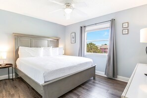 A bedroom with a bed, nightstand, lamp, ceiling fan, window with curtains, and a view of buildings outside. - Villas on the Gulf A4 | Steps to Beach | Pool (Gulf Breeze)