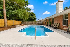A rectangular swimming pool with a ladder, surrounded by a concrete deck, a wooden fence, and a brick building with windows and outdoor seating. - Private Pool Game Room 8 Min to Perdido Key (Pensacola)