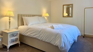 A bedroom with a bed, two bedside lamps, a nightstand, a mirror, and a chandelier. - Chepstow 4BR Retreat | Gated, Garden & Parking (Chepstow)