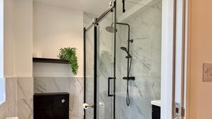 A modern bathroom with a glass-enclosed shower, a toilet, a trash bin, and a shelf with a plant. - Chepstow 4BR Retreat | Gated, Garden & Parking (Chepstow)