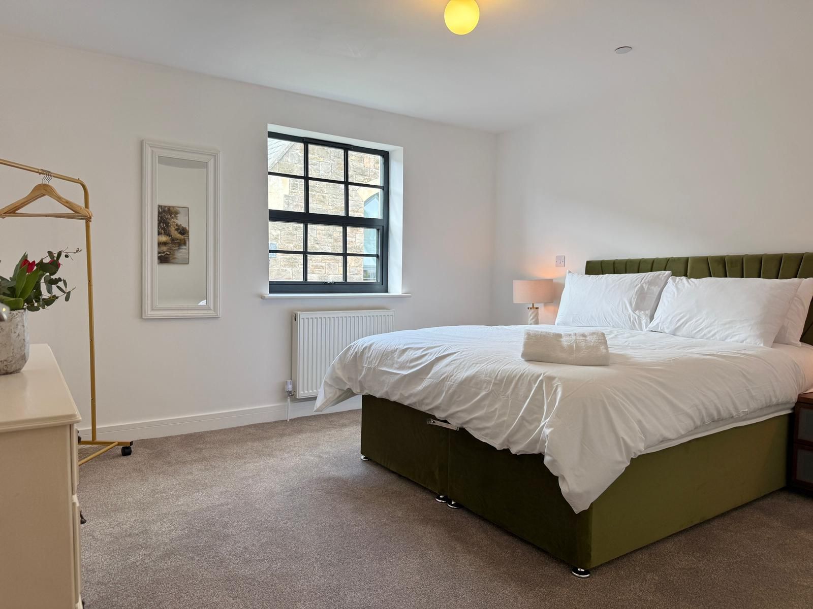 Une chambre à coucher avec un lit, un radiateur, une fenêtre, une table de chevet avec une lampe et une étagère murale agrémentée d’une plante.