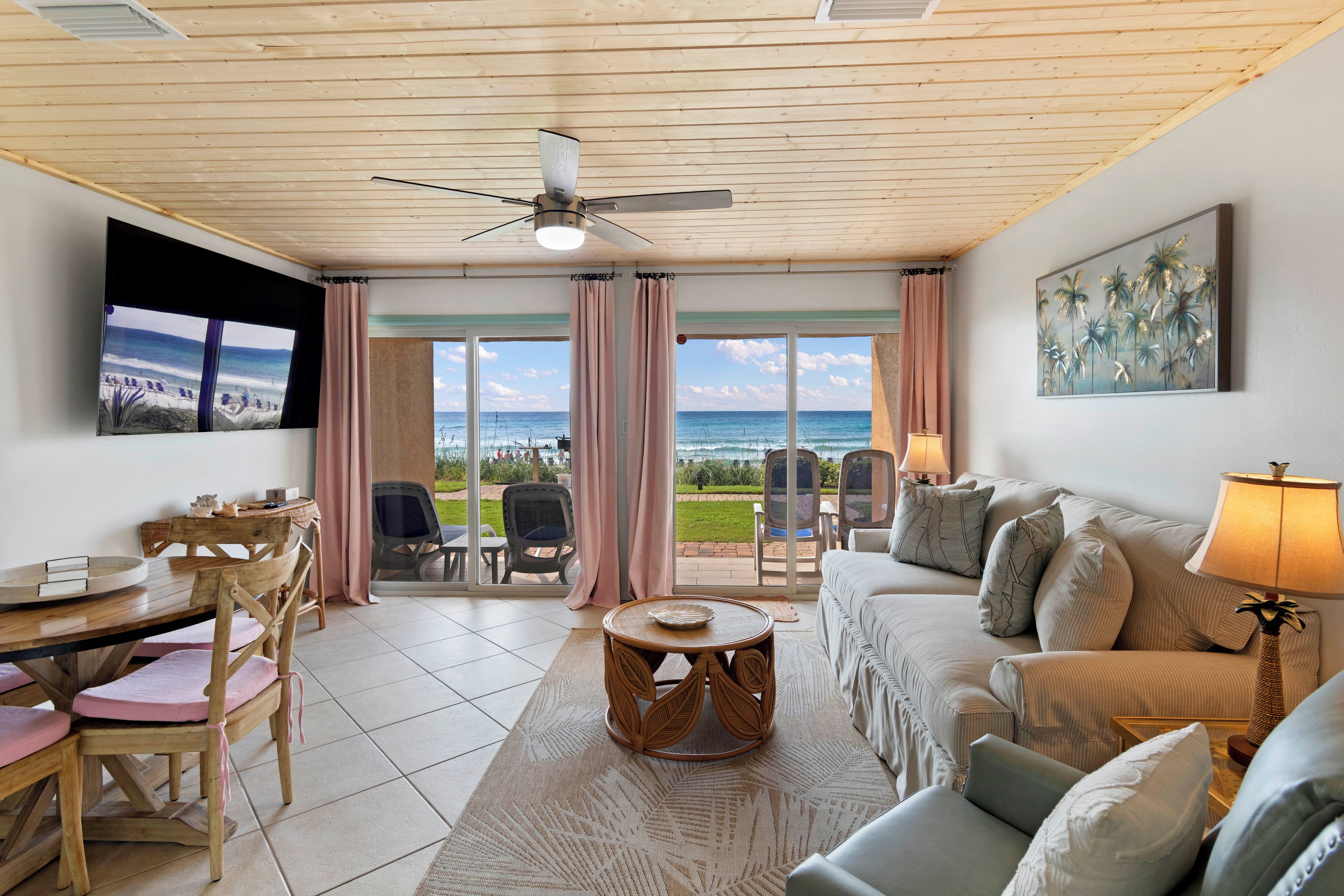 A room with a ceiling fan, a television, a sofa, a table, chairs, and a view of the ocean through large windows.