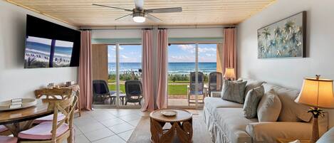 A room with a ceiling fan, a television, a sofa, a table, chairs, and a view of the ocean through large windows.