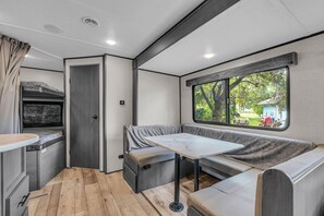 A compact RV interior with a kitchenette, a dining area, and a window with a view of greenery.