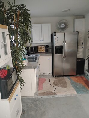 Una cocina compacta con armarios blancos, un refrigerador de acero inoxidable, horno empotrado y microondas. Hay un lavamanos pequeño y un ventilador en la pared.