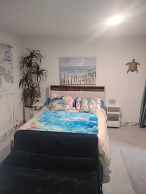 Un dormitorio con una cama, una mesita de noche, una obra de arte en la pared, una planta en maceta y una pequeña mesa.