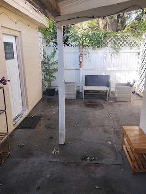 Ein überdachter Patio-Bereich mit Betonboden, einer Holzbank, einem Blumentopf und einem weißen Zaun.