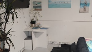 Ett rum med ett vitt skrivbord, en svart soffa, en tv som är monterad på väggen och en krukväxt.