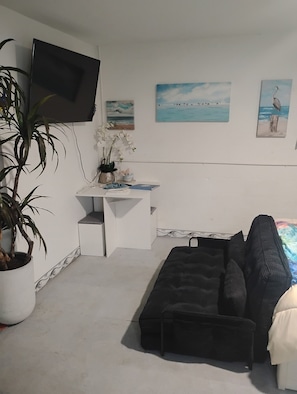 Una habitación con un escritorio blanco, un sofá negro, un televisor montado en la pared y una planta en maceta.