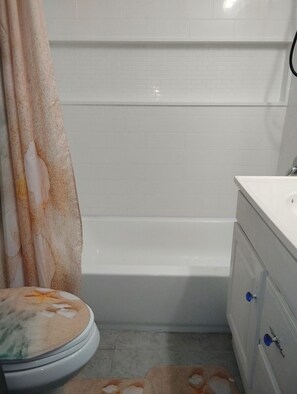 Un baño con una bañera blanca, un inodoro con cubierta floral y un tocador con espejo y pomo azul.